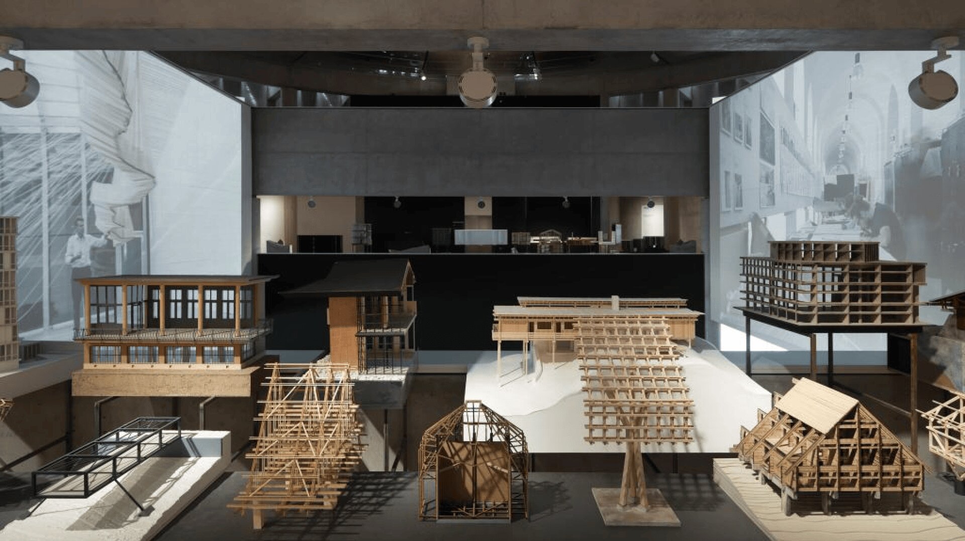 Eine Ausstellung von architektonischen Holzmodellen, die auf Plattformen in einem modernen Ausstellungsraum angeordnet sind, mit großen Schwarz-Weiß-Architekturbildern, die auf die umliegenden Wände projiziert werden.