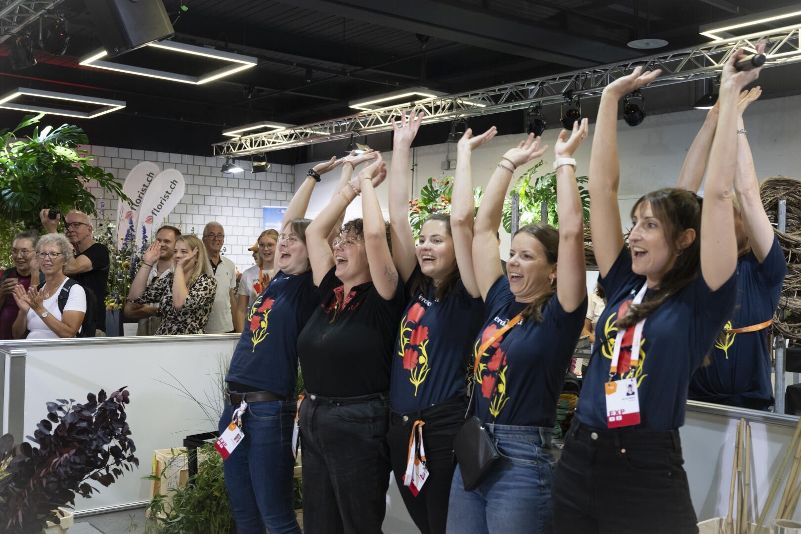 Eine Gruppe von Frauen in farblich abgestimmten Hemden steht mit erhobenen Armen lächelnd und jubelnd bei einer Indoor-Veranstaltung. Auch die Menschen im Hintergrund applaudieren und schauen zu, umgeben von Pflanzen und Dekorationen.