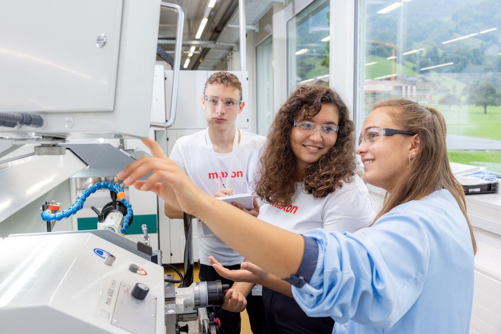 Drei junge Leute mit Schutzbrillen stehen an einer Maschine in einem hellen Labor. Eine Person gestikuliert, während sie etwas erklärt, und die beiden anderen hören aufmerksam zu und machen sich Notizen. Große Fenster zeigen eine grüne Landschaft draußen.