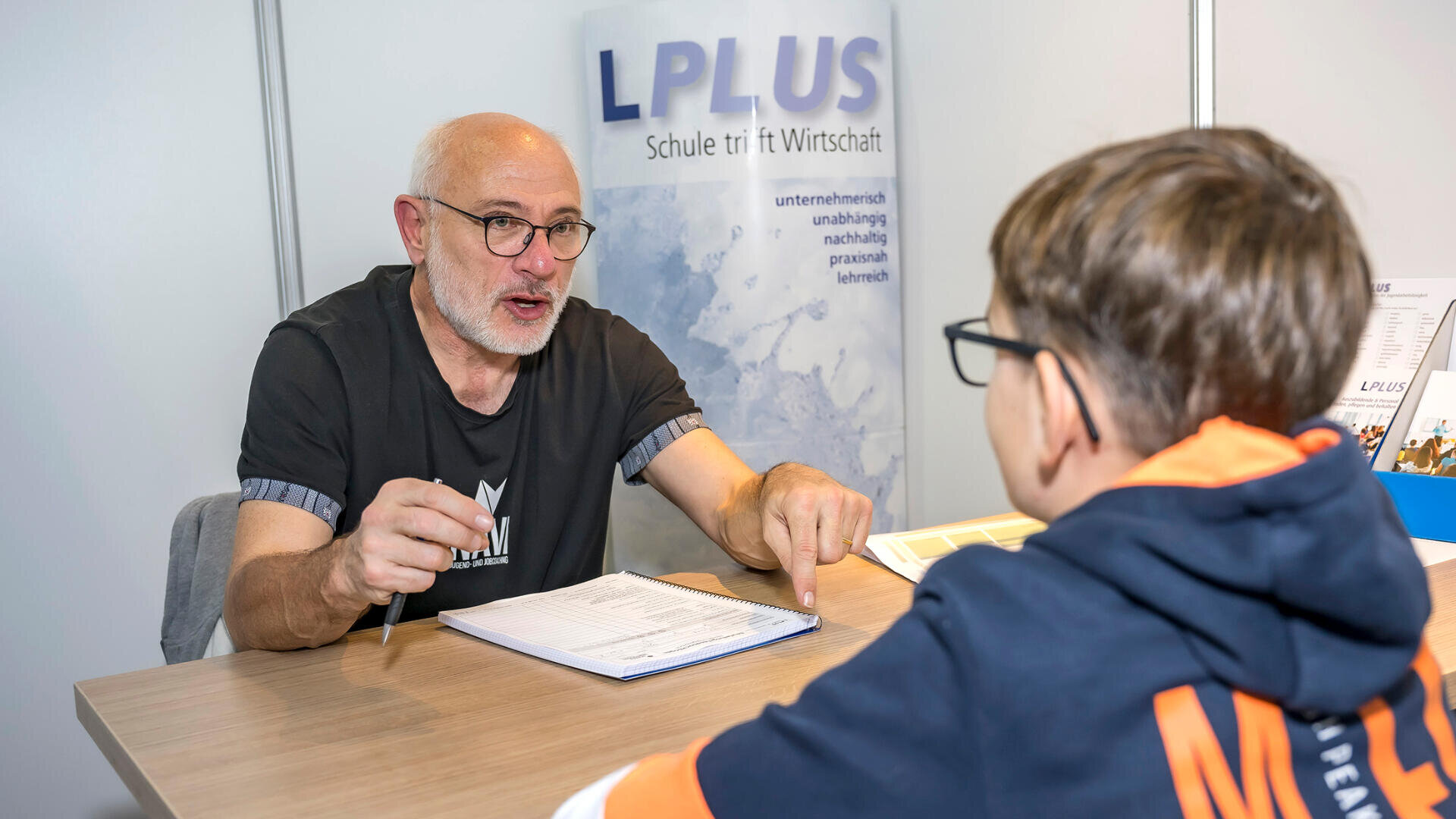 Ein älterer Mann mit Brille unterhält sich mit einem Jungen, der einen Kapuzenpulli trägt, über einen Tisch hinweg, hält einen Stift in der Hand und zeigt auf Dokumente, während im Hintergrund ein Schild der LPLUS Schule zu sehen ist.