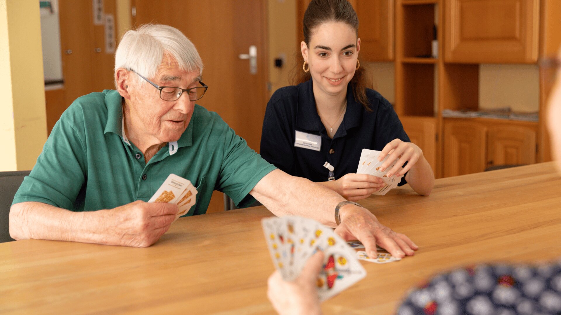 Ein älterer Mann und eine junge Frau sitzen an einem Holztisch und spielen mit einer anderen Person Karten. Der Mann macht einen Zug, während die anderen ihre Karten halten, lächeln und das Spiel gemeinsam genießen.