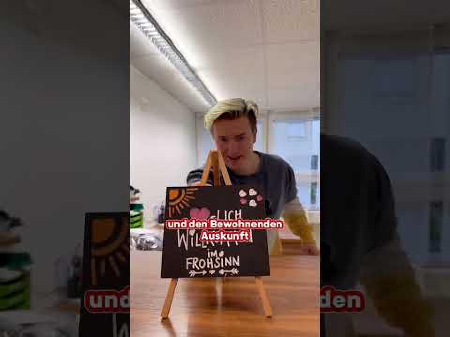 Eine Person mit blondem Haar steht hinter einer kleinen Staffelei, die ein schwarzes Schild mit bunten Schriftzügen und Zeichnungen zeigt. Der rote deutsche Text darüber lautet "und den Bewohnenden Auskunft". Der Schauplatz scheint ein Innenraum zu sein.