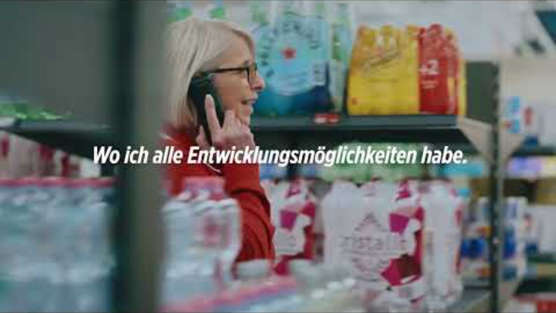 Eine Frau mit grauen Haaren und Brille, die einen roten Pullover trägt, telefoniert, während sie im Gang eines Lebensmittelgeschäfts steht. Der deutsche Text auf dem Bild lautet: "Wo ich alle Entwicklungsmöglichkeiten habe.