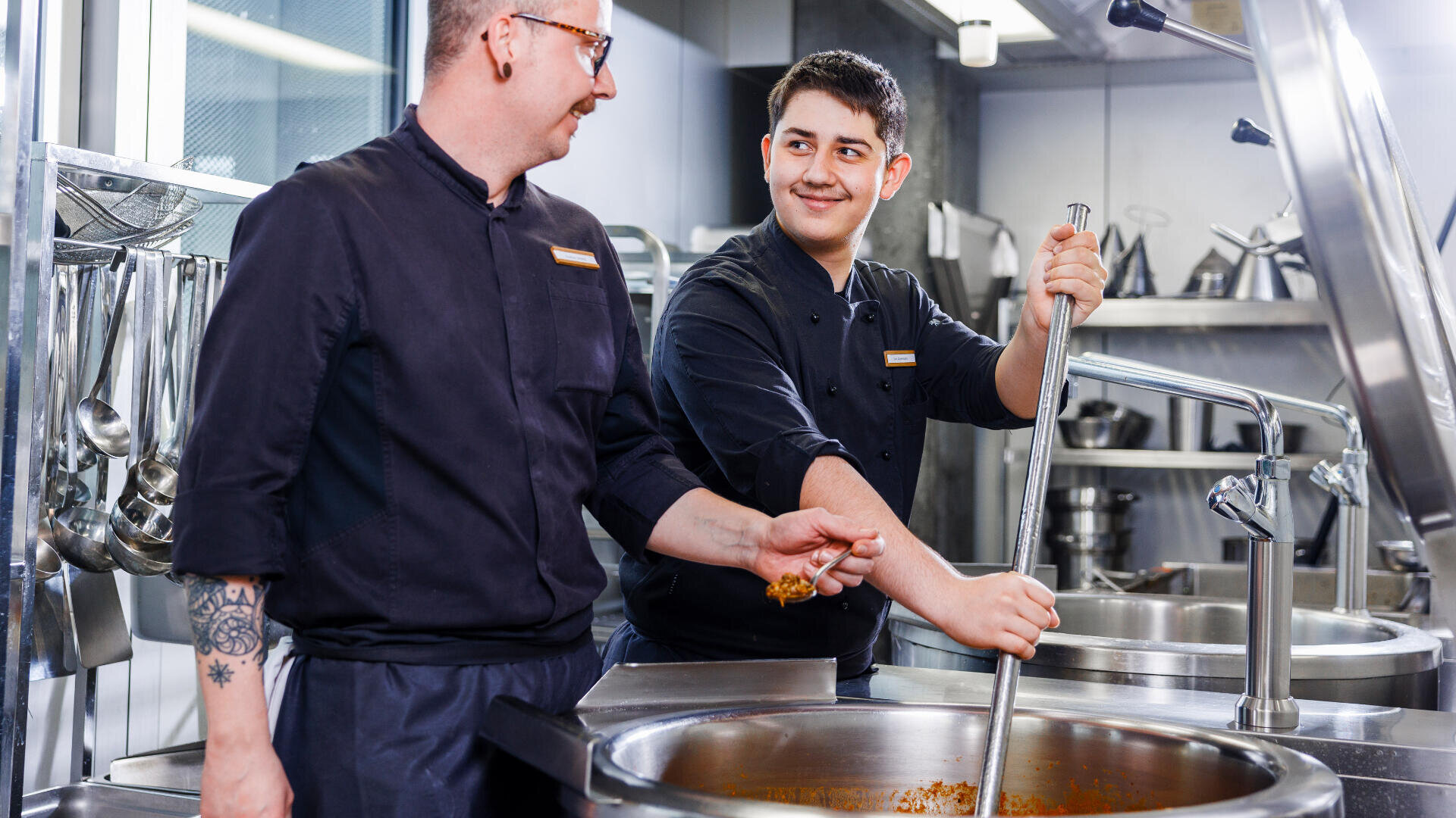 Zwei Köche in schwarzen Uniformen arbeiten zusammen in einer Großküche, rühren einen großen Topf mit Essen um und lächeln sich an. Im Hintergrund sind Küchenutensilien und Geräte aus Edelstahl zu sehen.