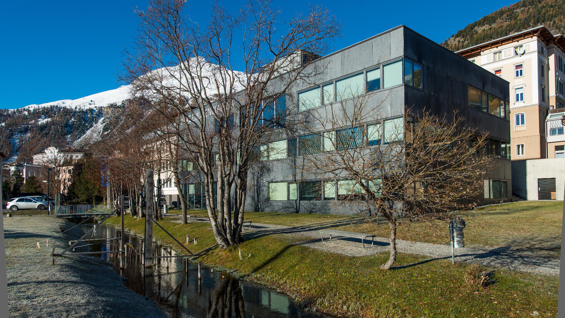 Ein modernes graues Gebäude mit großen Fenstern steht neben einem kleinen Bach und blattlosen Bäumen, mit schneebedeckten Bergen und einem klaren blauen Himmel im Hintergrund.