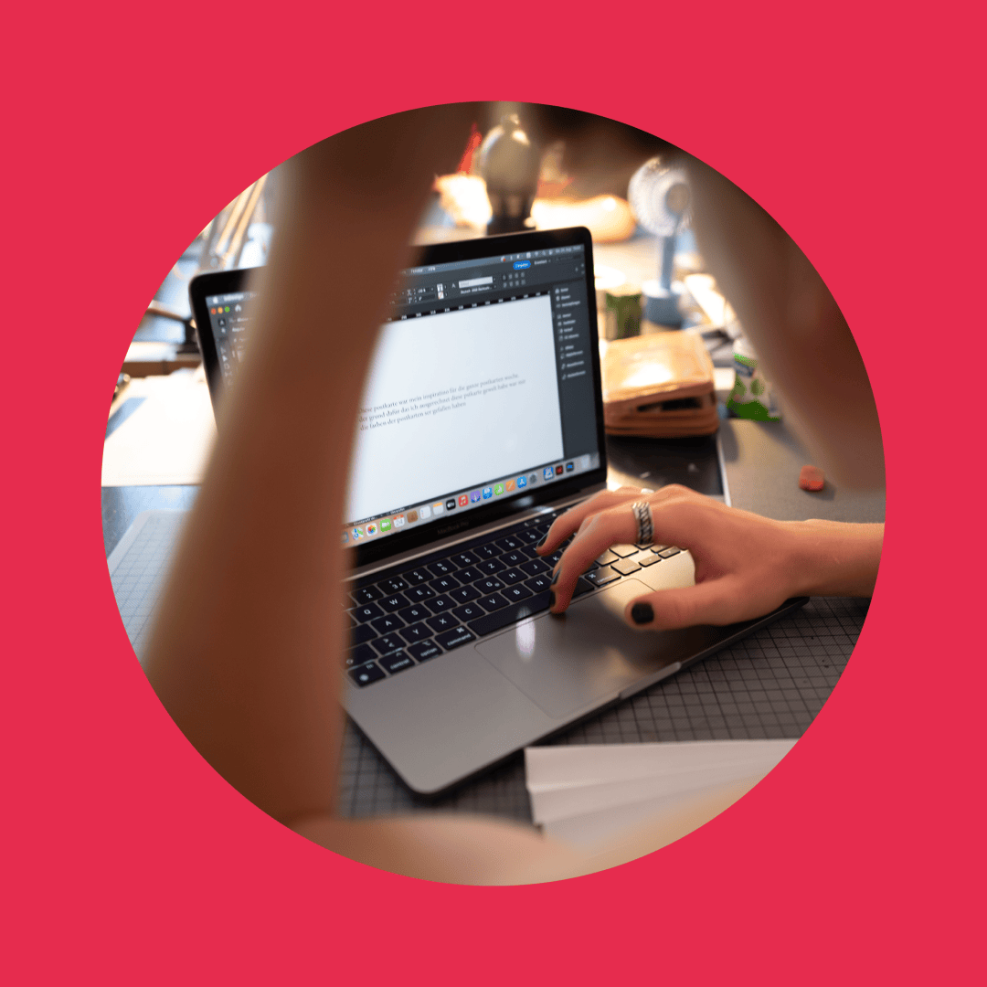 Eine Person mit lackierten Nägeln tippt auf einem Laptop und bearbeitet ein Dokument. Die Szene wird durch einen kreisförmigen Rahmen mit rosa Hintergrund fokussiert, und im Hintergrund ist ein unordentlicher Schreibtisch zu sehen.