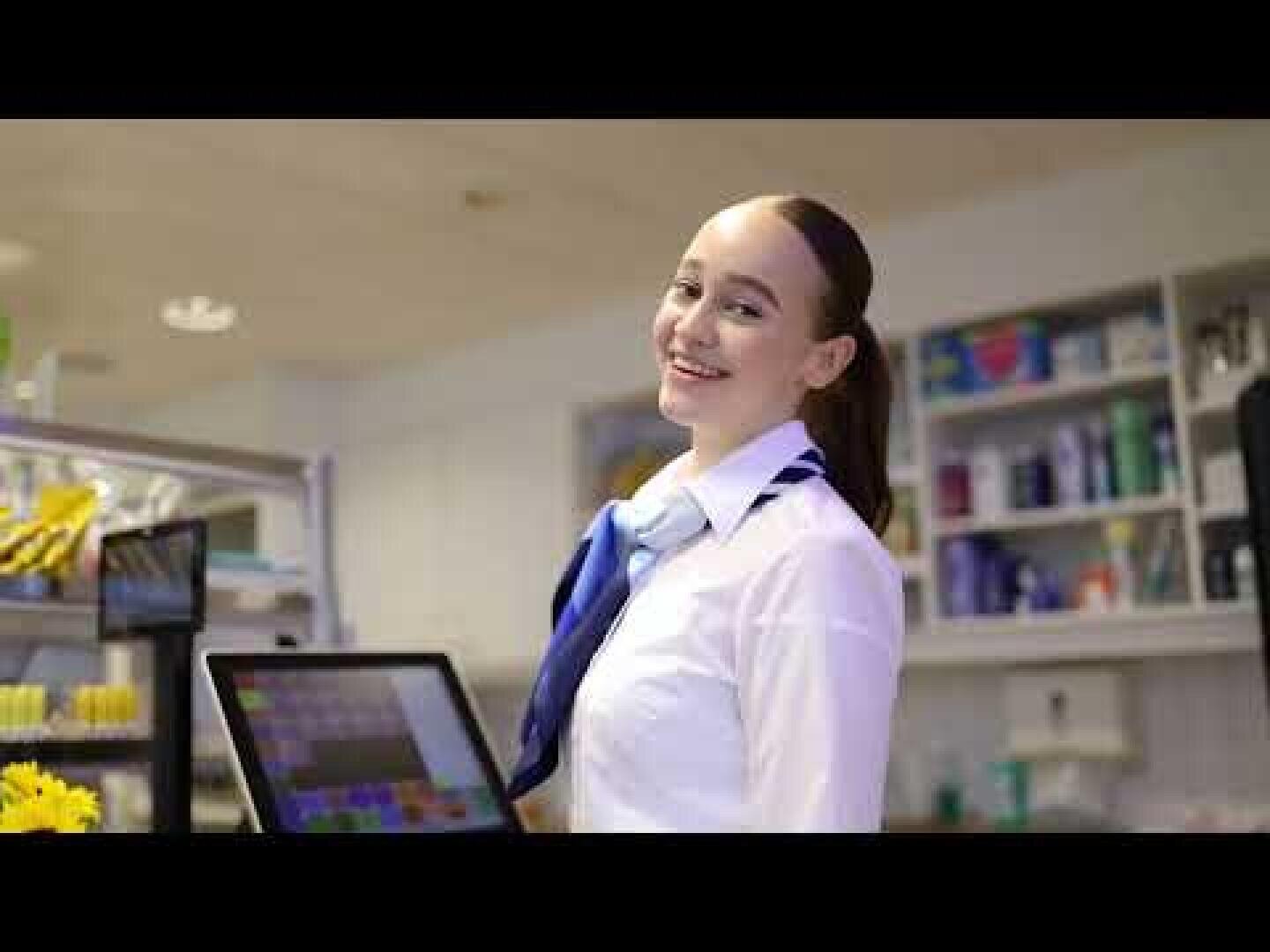 Eine lächelnde Frau mit weißem Hemd und blauem Schal steht hinter einer Ladentheke in einem Geschäft oder Café, mit Regalen mit verschiedenen Produkten im Hintergrund. Sie befindet sich neben einer Touchscreen-Kasse.