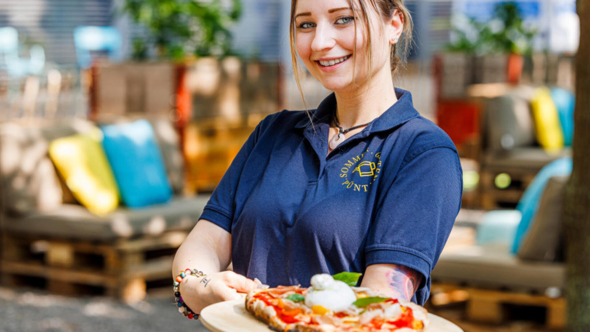 Ein lächelnder Kellner in einem marineblauen Polohemd präsentiert eine frisch zubereitete Pizza auf einem Holzbrett in einem Essbereich im Freien mit bunten Kissen und Holzstühlen im Hintergrund.