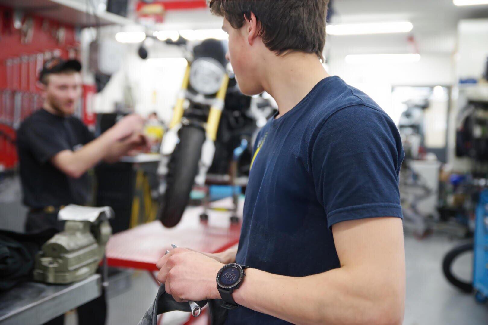 Ein junger Mann mit einem marineblauen Hemd und einer Smartwatch steht in einer Motorradwerkstatt und hält ein Werkzeug in der Hand, während eine andere Person neben einem Motorrad auf einer Hebebühne im Hintergrund gestikuliert und spricht.