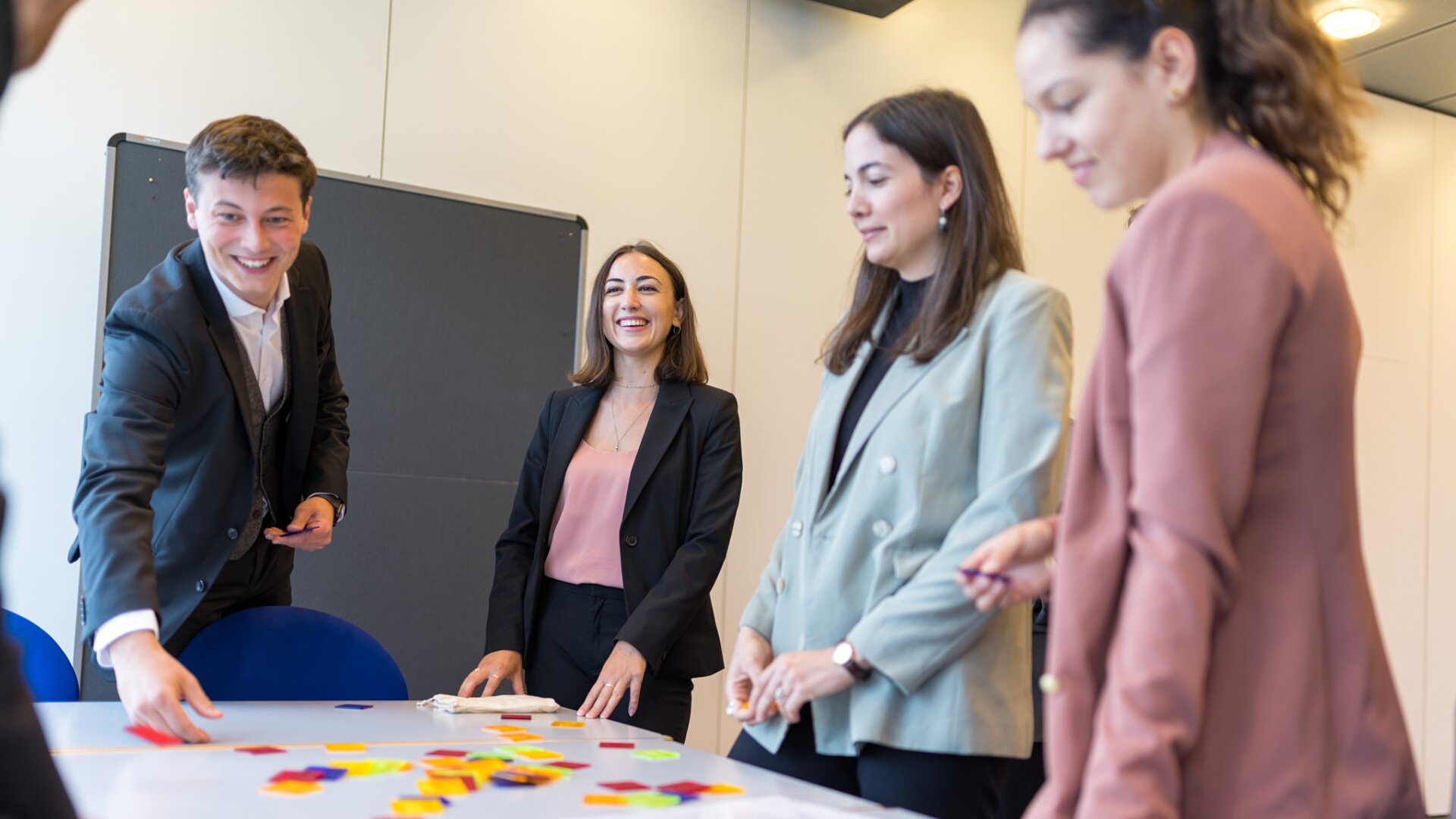 Vier Personen in Geschäftskleidung stehen um einen Tisch, der mit bunten Formen bedeckt ist, lächeln und interagieren, was auf eine gemeinschaftliche oder teambildende Aktivität in einer modernen Büroumgebung hindeutet.