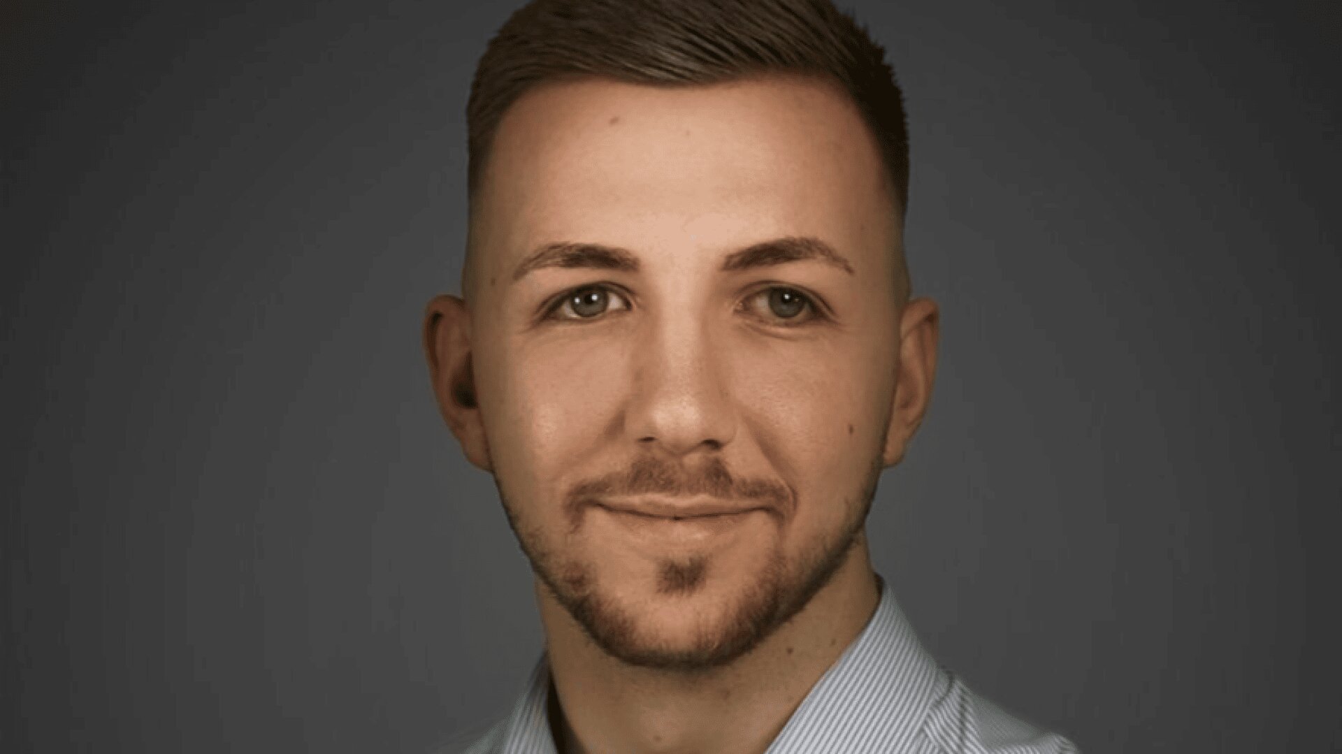 Ein junger Mann mit kurzen braunen Haaren, ordentlich gestutztem Bart und heller Haut lächelt leicht. Er trägt ein helles, gestreiftes Hemd mit Kragen und steht vor einem schlichten dunklen Hintergrund.