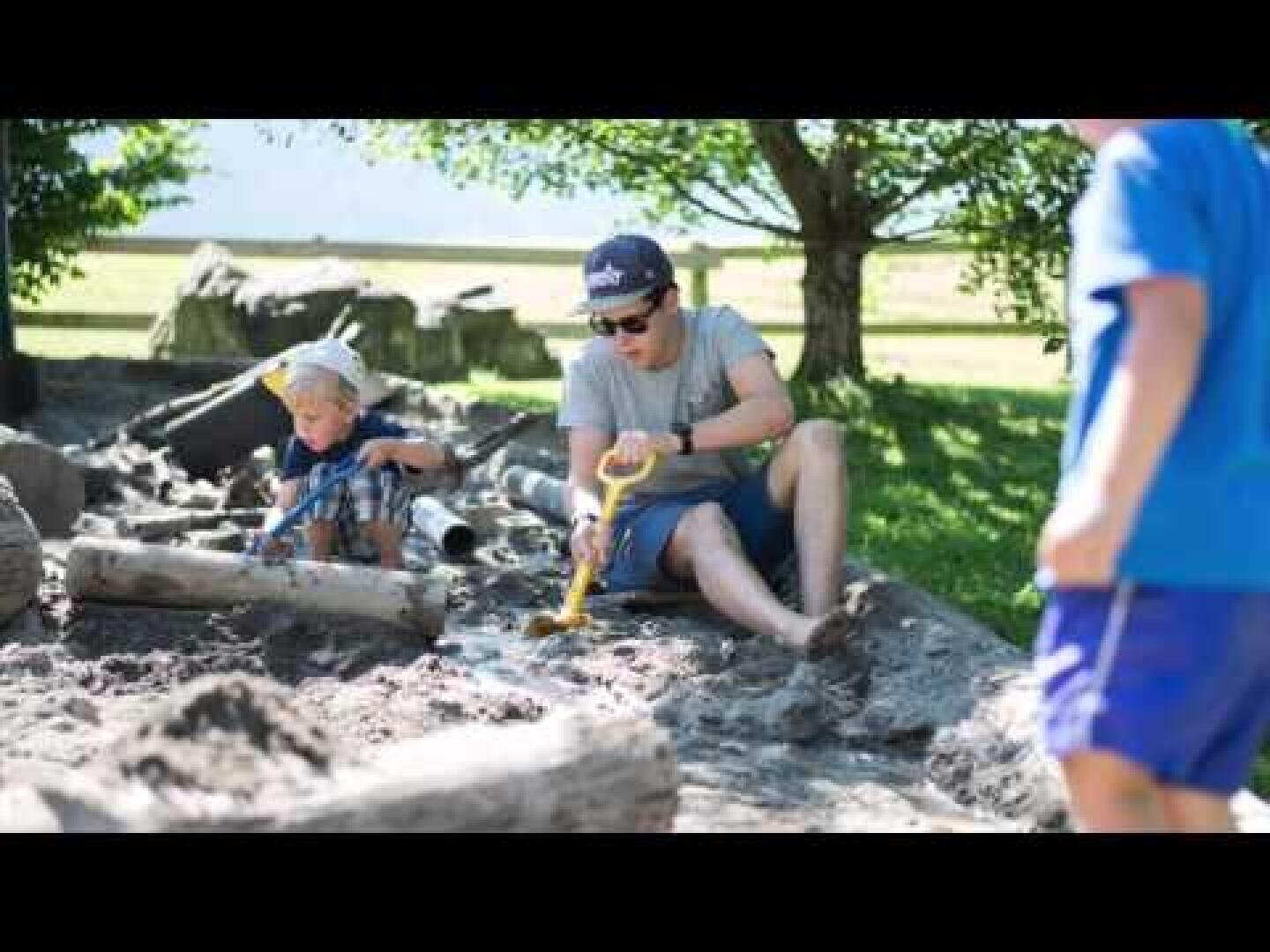 Ein Erwachsener und ein Kind spielen mit Spielzeugschaufeln in einem sandigen Außenbereich, der von Baumstämmen umgeben ist. Ein anderes Kind in blauen Shorts steht in der Nähe. An einem sonnigen Tag sind im Hintergrund Bäume und Gras zu sehen.