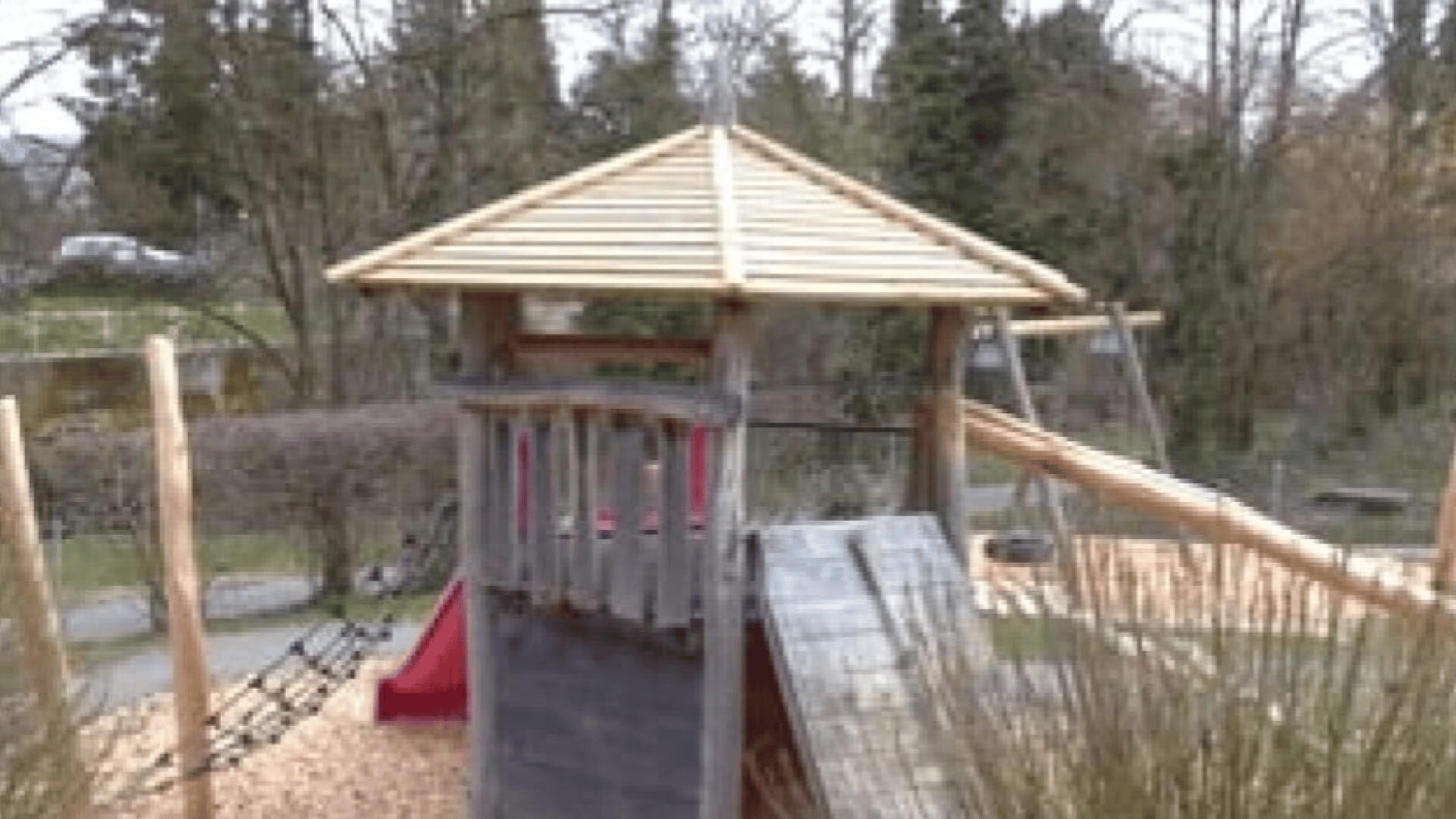 Ein Spielplatz aus Holz mit Spitzdach, Kletterwand, Rutsche und Seilen, umgeben von Bäumen und Sträuchern in einer Parklandschaft.