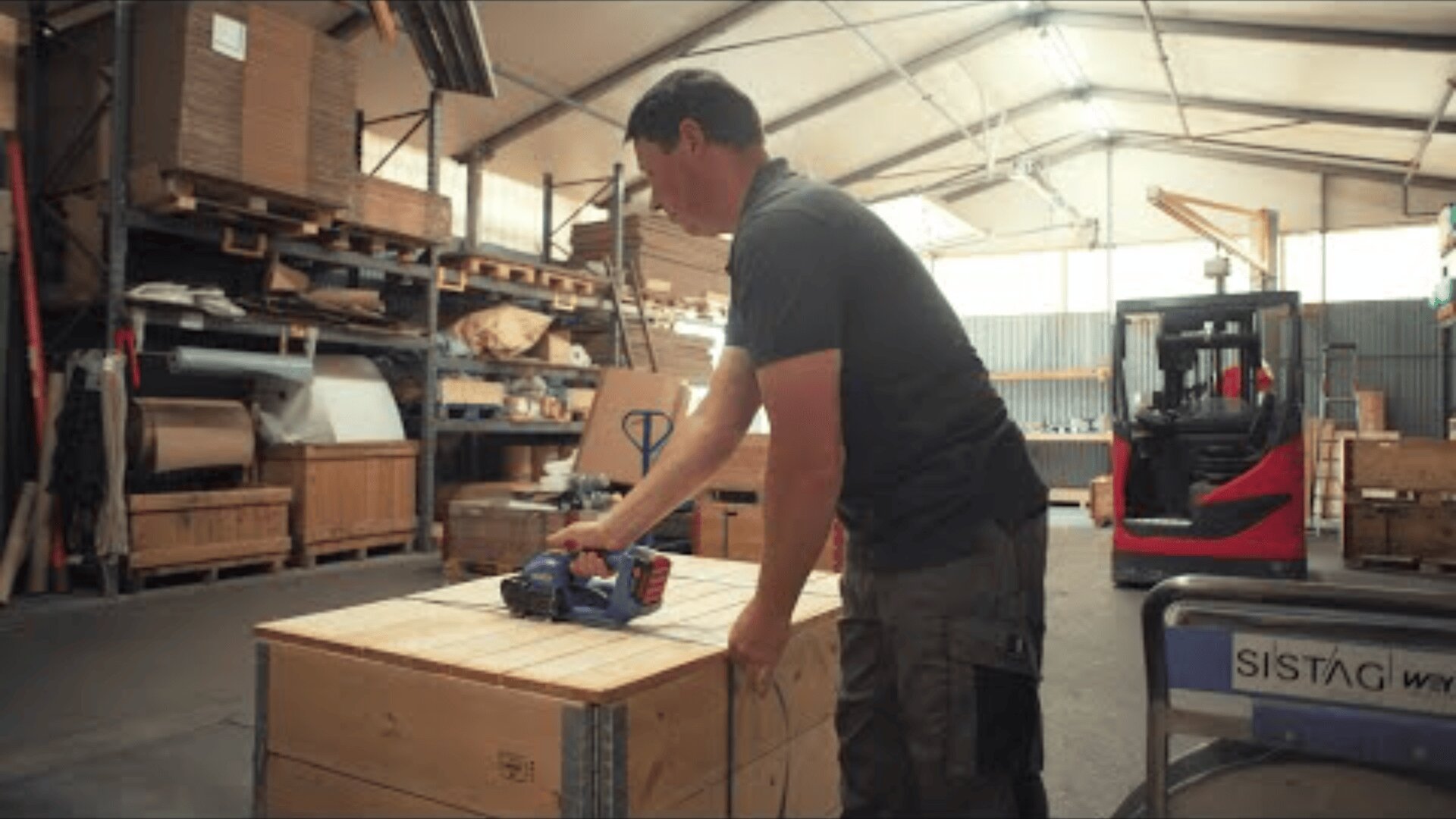 Eine Person in einer Werkstatt verwendet ein handgehaltenes Werkzeug, um eine große Holzkiste zu umreifen. Im Hintergrund sind Regale mit Materialien und ein roter Gabelstapler zu sehen.
