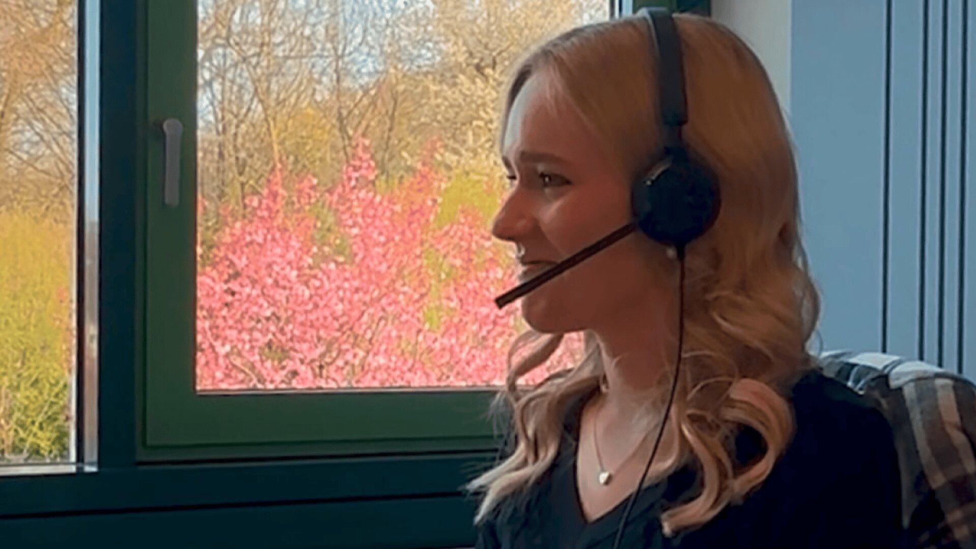 Eine Frau mit blondem Haar und Kopfhörer sitzt am Fenster und lächelt. Außerhalb des Fensters sind Bäume mit rosa und grünem Laub zu sehen, was auf den Frühling hindeutet.