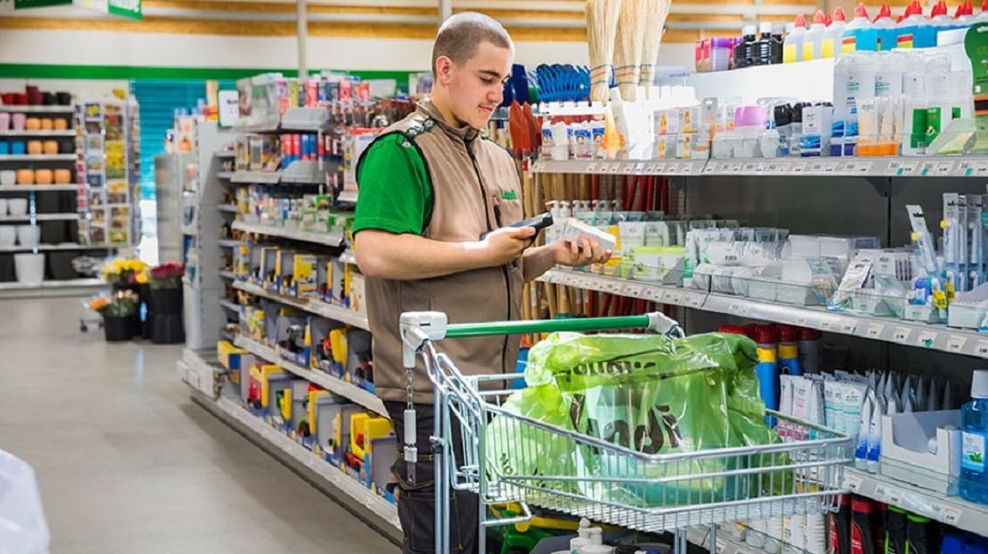 Ein Angestellter in einer braun-grünen Uniform durchsucht ein Regal, während er einen Einkaufswagen in einem gut beleuchteten Geschäft mit verschiedenen Produkten füllt.