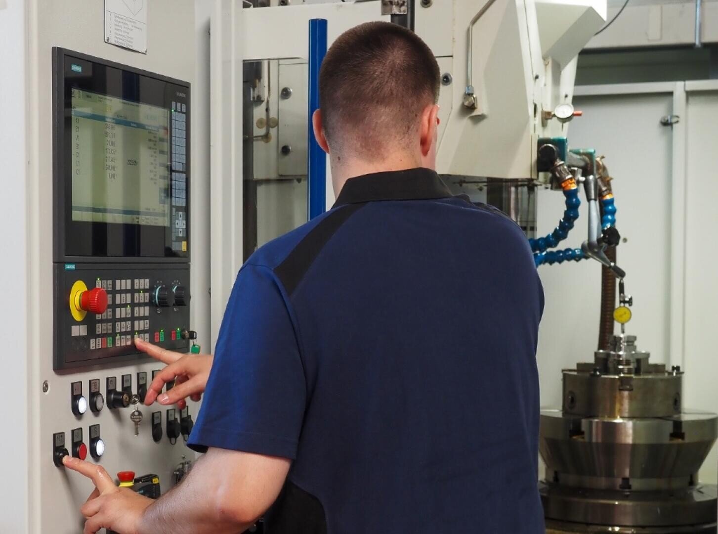 Eine Person, die eine CNC-Maschine bedient, drückt Tasten auf einem Bedienfeld mit einem Bildschirm in einer industriellen Umgebung, in der Maschinen im Hintergrund zu sehen sind.