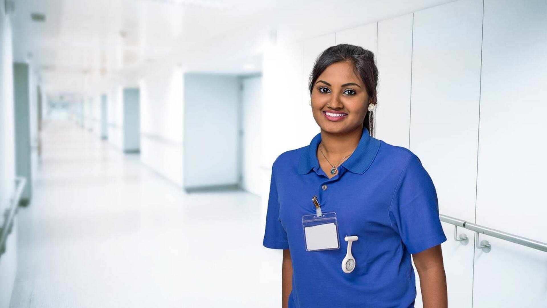 Ein lächelnder Mitarbeiter des Gesundheitswesens mit dunklem Haar, der eine blaue Uniform und einen Ausweis trägt, steht in einem hellen, modernen Krankenhauskorridor.