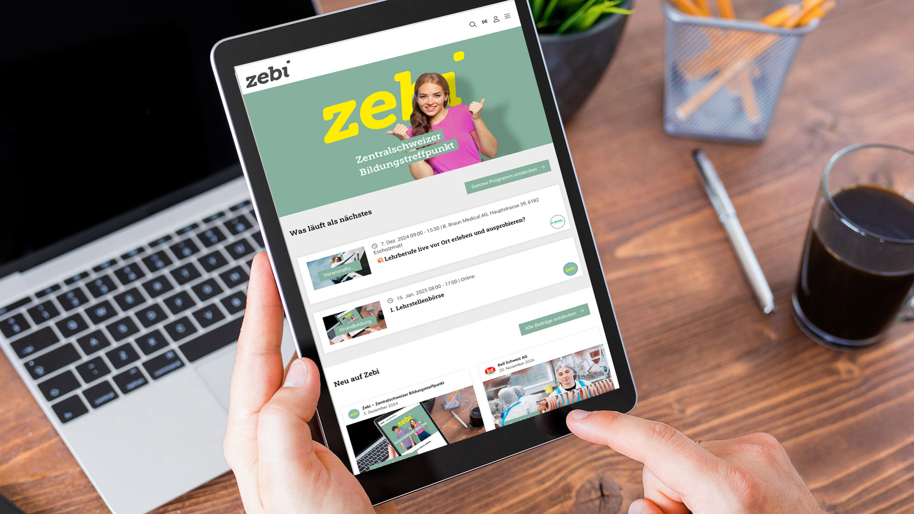 Person hält ein Tablet, auf dem die Zebi-Website auf Deutsch angezeigt wird, mit einer lächelnden Frau auf der Homepage, daneben ein Laptop, ein Stifthalter und ein Glas mit dunkler Flüssigkeit auf einem Holzschreibtisch.