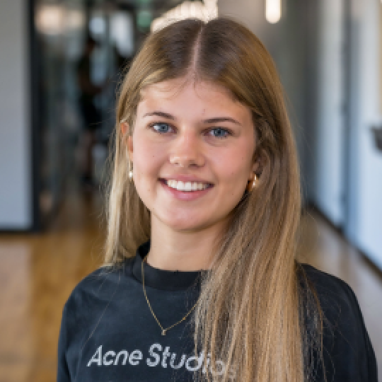 Eine junge Frau mit langen blonden Haaren lächelt, während sie ein schwarzes Sweatshirt von Acne Studios trägt.