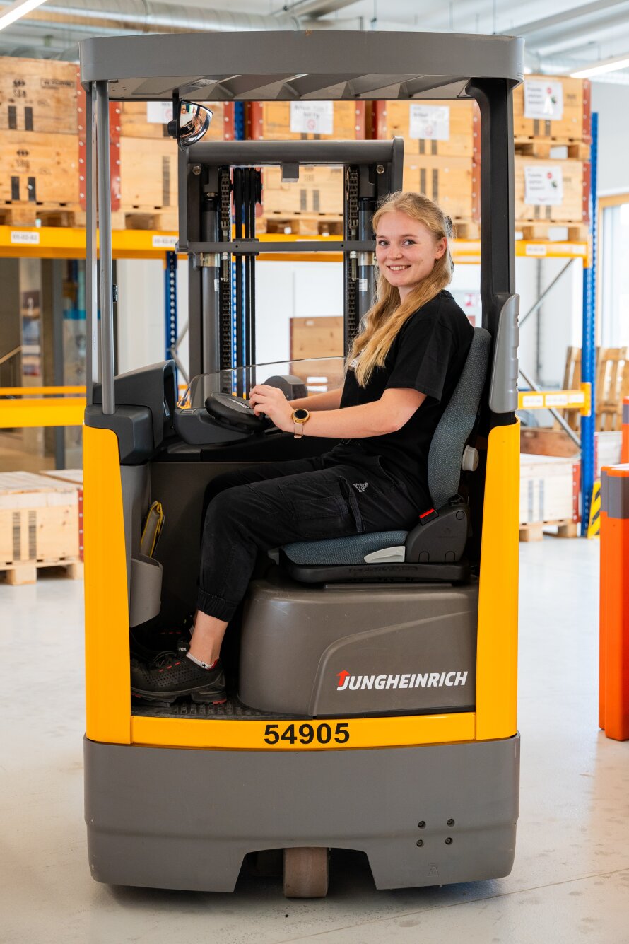 Eine junge Frau lächelt, während sie sitzend einen gelben Jungheinrich-Gabelstapler in einem Lagerhaus mit Regalen und Holzkisten im Hintergrund bedient.