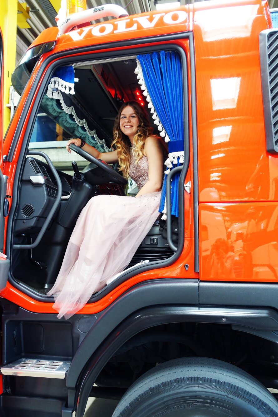 Eine junge Frau in einem rosafarbenen Kleid sitzt lächelnd auf dem Fahrersitz eines knallroten Volvo-Trucks, der mit blauen Vorhängen und weißer Spitzenverzierung geschmückt ist.