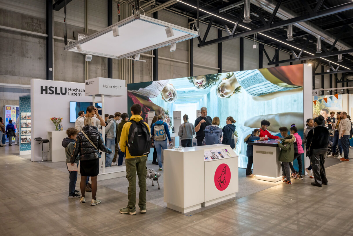 Besucher treffen sich an einem Bildungsstand mit der Beschilderung der HSLU Hochschule Luzern. Grosse Bildschirme zeigen Bilder über den Ständen, und Besucher interagieren mit Exponaten und Mitarbeitern in einer geräumigen, modernen Halle.