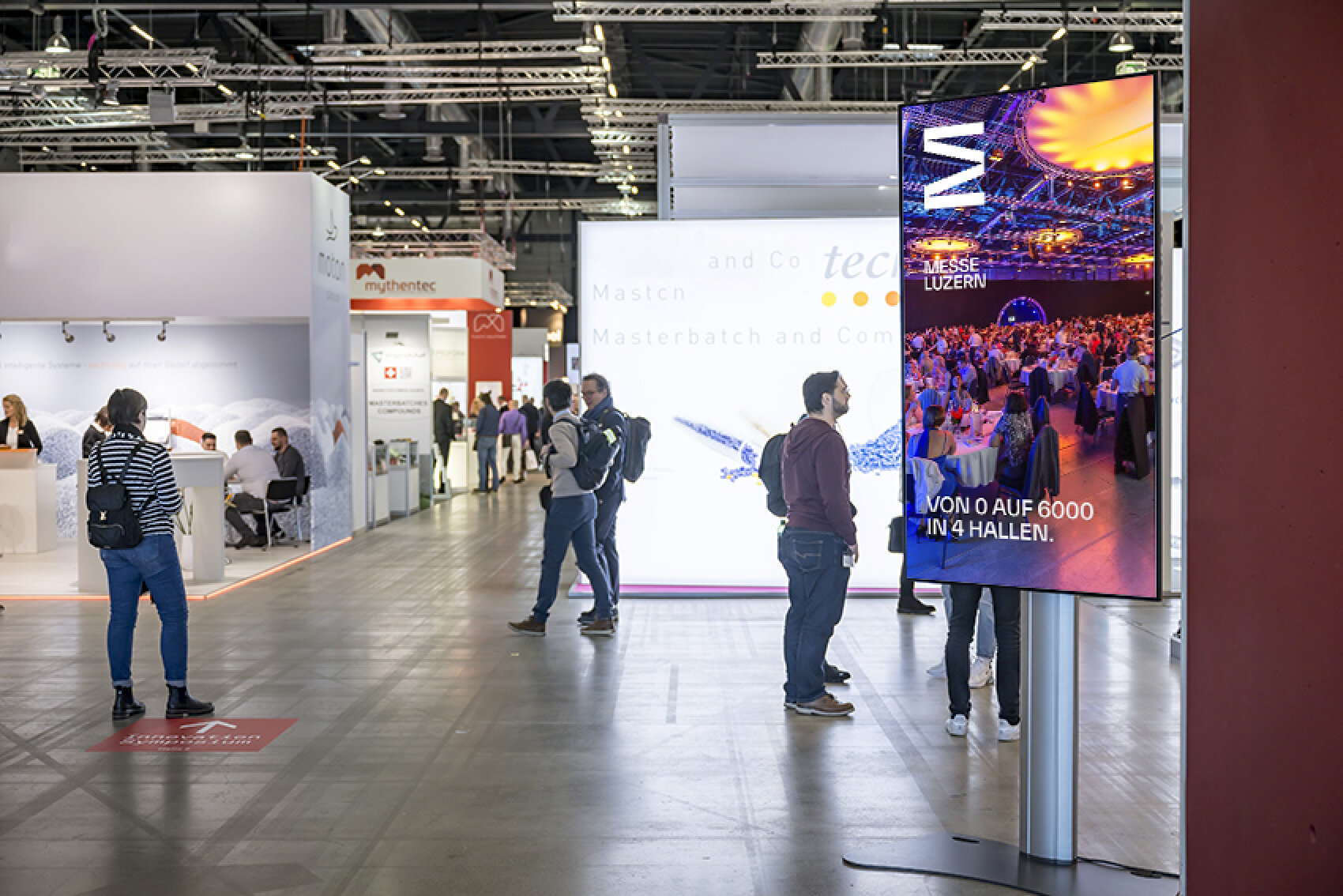 Ein grosser digitaler Bildschirm in einer Messehalle zeigt Werbung f&uuml;r die Messe Luzern mit dem Text 'Von 0 auf 6000 in 4 Hallen'. Im Hintergrund sind Messest&auml;nde und Besucher sichtbar.
