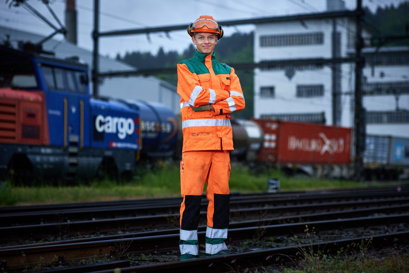 Ein Eisenbahner, der orangefarbene und grüne Warnschutzkleidung und einen Helm trägt, steht mit verschränkten Armen auf den Gleisen, mit Güterzügen und Industriegebäuden im Hintergrund.