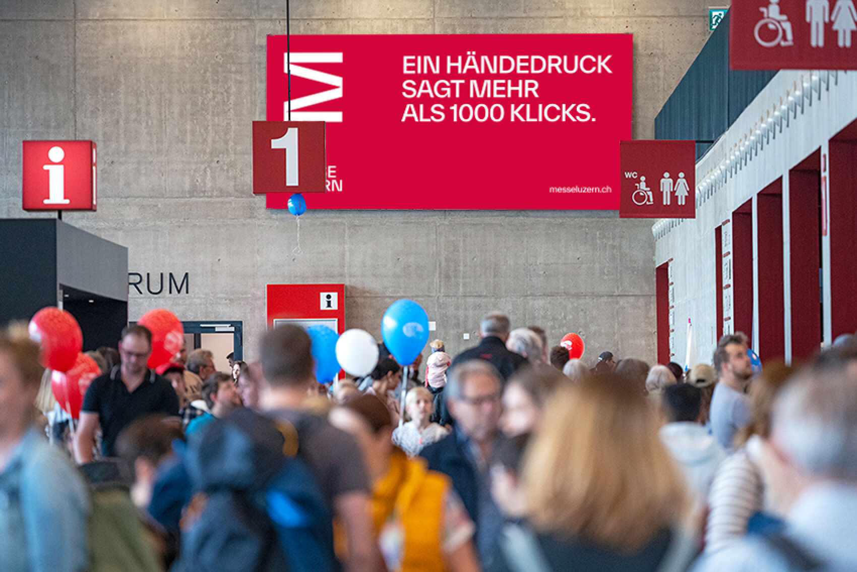 Ein grosses rotes Banner in einer Messehalle mit der Aufschrift 'Ein H&auml;ndedruck sagt mehr als 1000 Klicks'. Darunter sind zahlreiche Messebesucher mit bunten Luftballons zu sehen.