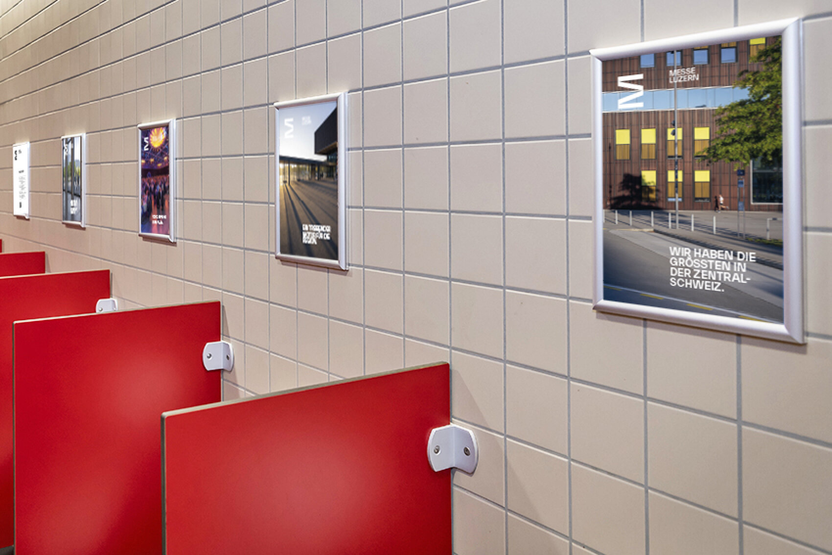 Eine Kachelwand in einer &ouml;ffentlichen Toilette, an der mehrere Poster angebracht sind. Jedes Poster zeigt unterschiedliche Werbeinhalte. Im unteren Bereich sind rote Trennw&auml;nde sichtbar.