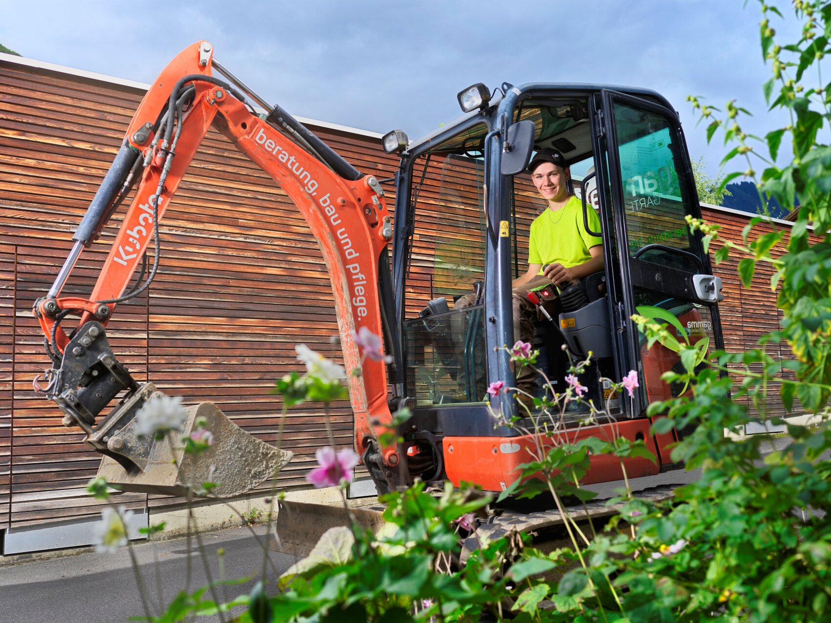 Eine Person in einem leuchtend gelben Hemd bedient einen kleinen orangefarbenen Bagger in der Nähe eines modernen Holzgebäudes, mit grünen Pflanzen und rosa Blumen im Vordergrund.